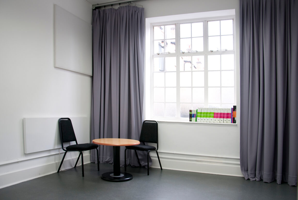 A room with a large window and purple curtains. The floor is grey rubber, and there is a table and two chairs in the corner of the room.