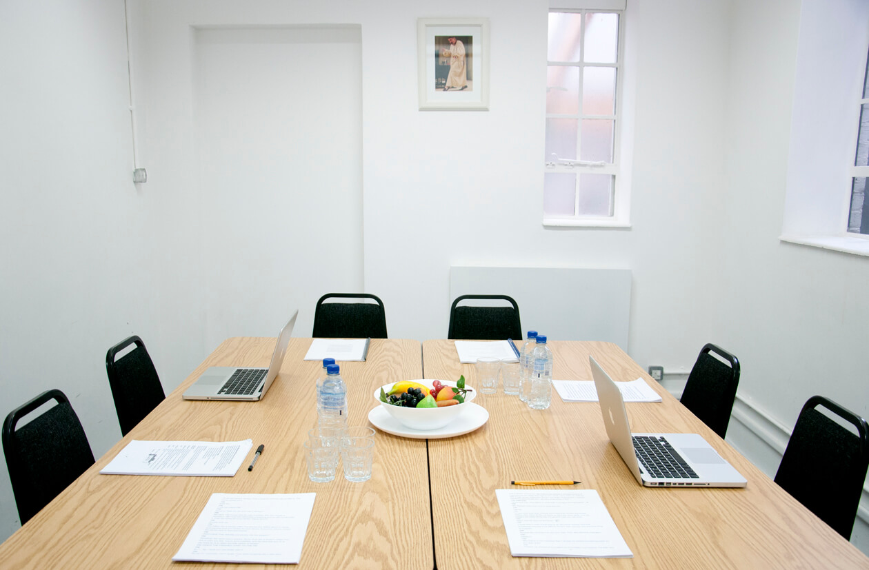 A meeting room with white walls and two windows. There is a large wooden table in the centre of the room, and six chairs around it. On the table is a bowl of fruit, bottles of water, laptops and meeting papers.