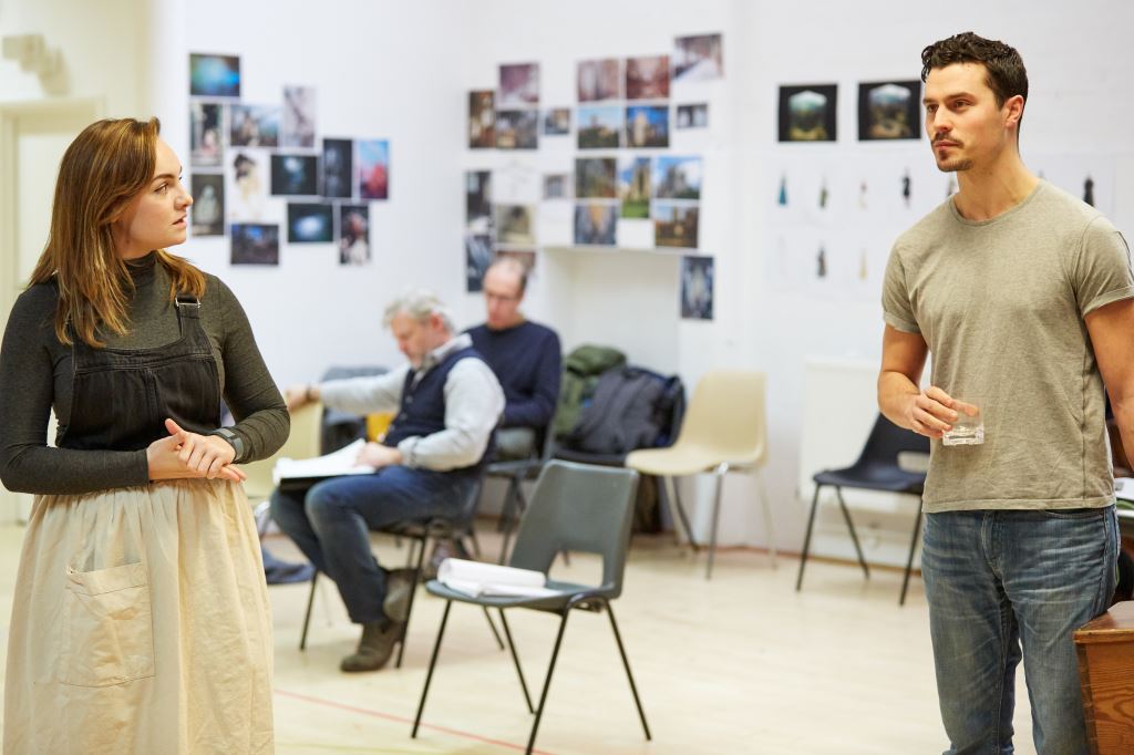 A rehearsal photograph of actors in a white studio. Two actors are in focus, in the foreground of the photo. In the background there are two other actors, out of focus, sat reading a script. The walls of the studio are covered in photographs.