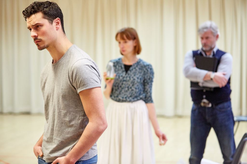 A rehearsal photograph of 3 actors in a white studio. One of the actors is in the foreground of the photo, in focus, with his hands in his pockets. The other actors are the background, out of focus.